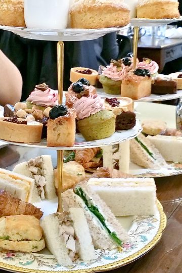 Two afternoon tea sets filled with mini desserts and sandwiches on a wooden tabletop.