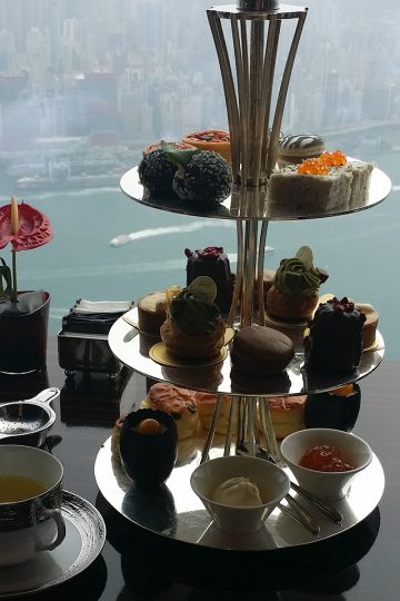 An afternoon tea set on a 3-tier silver tray in front of a window overlooking Hong Kong harbor.
