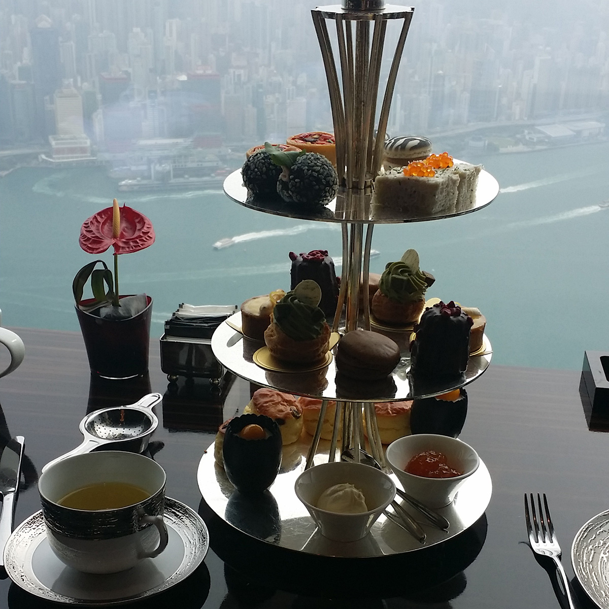 An afternoon tea set on a 3-tier silver tray in front of a window overlooking Hong Kong harbor.
