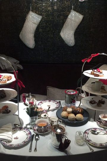 Two Christmas themed afternoon tea sets served on a traditional 3-tier tray place on a table in front of 2 Christmas stockings.
