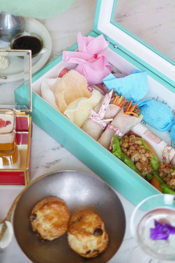 A tiffany blue jewel box is holding little afternoon tea treats laid out with other tea snacks like scones and pudding and champagne on a marble table.